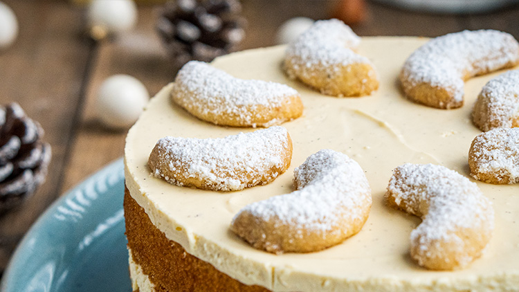 Runde Torte mit heller Creme und Vanillekipferln, die mit Puderzucker bestreut sind, auf blauem Teller dekoriert.