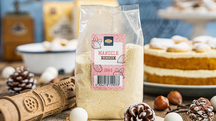 Plastikverpackung mit gemahlenen Mandeln, 200 g, steht auf einem Tisch mit Nüssen, Holzrolle und Torte im Hintergrund.