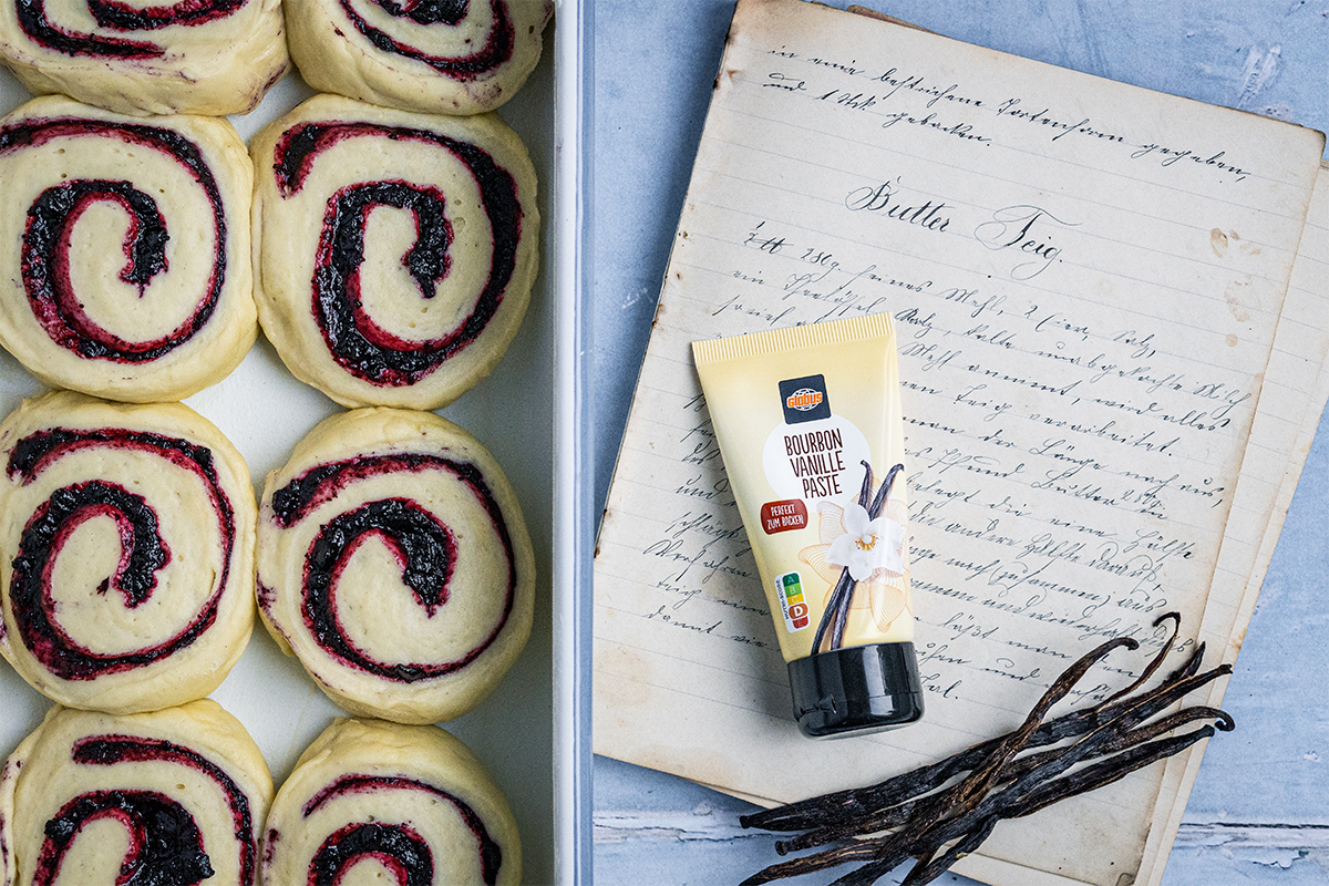 Sechs ungebackene Hefeschnecken mit roter Füllung, daneben ein Rezeptbuch, Vanilleschoten und eine Tube Bourbon Vanille Paste von GLOBUS.