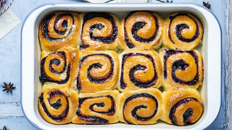 Zwölf goldbraune Hefeschnecken mit dunkler Füllung liegen in einer rechteckigen weißen Auflaufform auf blauem Untergrund.