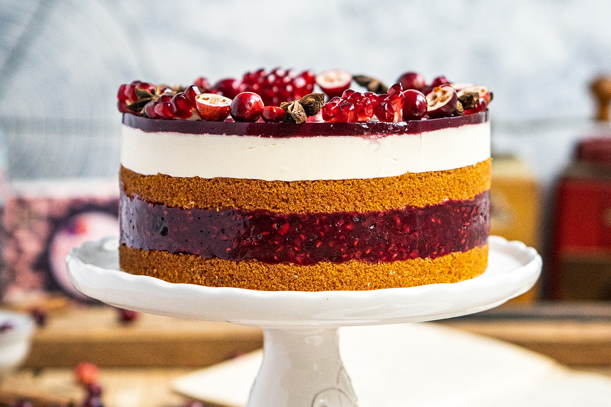Eine mehrschichtige Torte mit hellem Boden, roter Fruchtfüllung, weißer Creme und Deko aus Granatapfelkernen steht auf einer Tortenplatte.