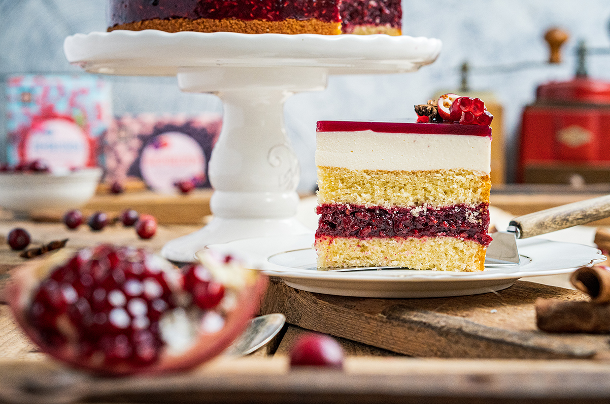Ein Stück Torte mit hellen und roten Schichten auf einem Teller, dekoriert mit roten Früchten, vor unscharfem Hintergrund.