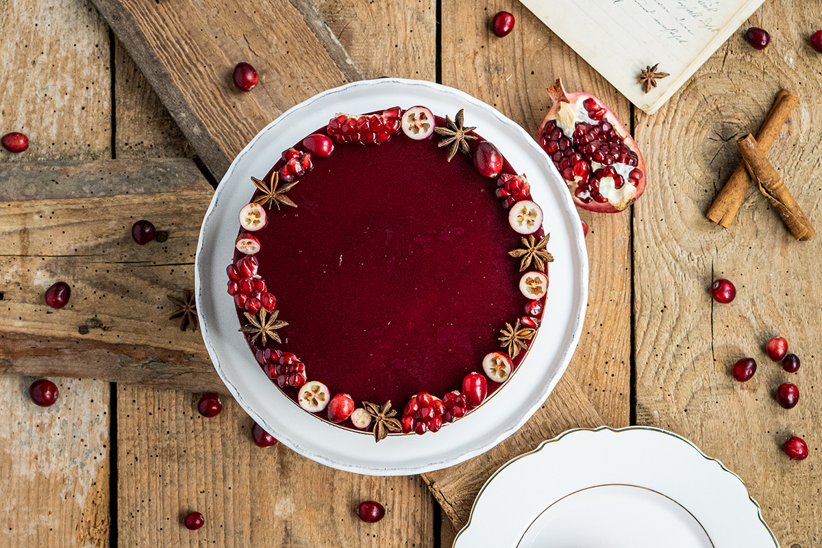 Runder Kuchen mit roter Glasur, verziert mit Sternanis, Granatapfelkernen, Cranberries und geschnittenen Beeren auf Holzuntergrund.