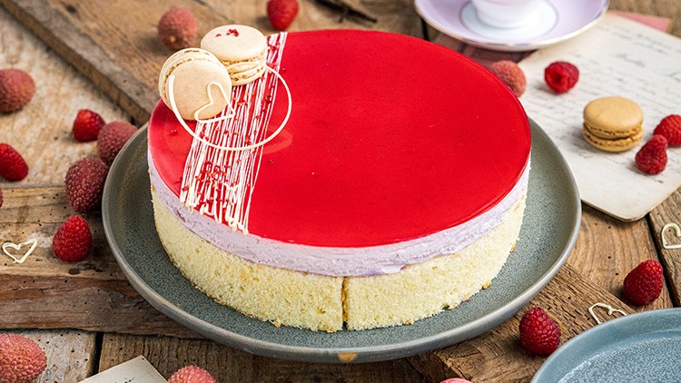 Torte mit roter Glasur, dekoriert mit Macarons und weißer Schokolade, auf grauem Teller mit frischen Himbeeren drumherum.