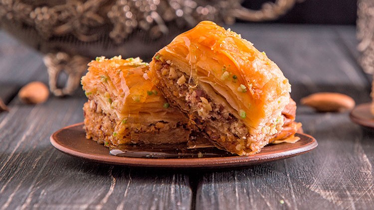 Zwei Stück Baklava mit Nüssen und Honig glasiert liegen auf einem braunen Teller auf rustikalem Holztisch.
