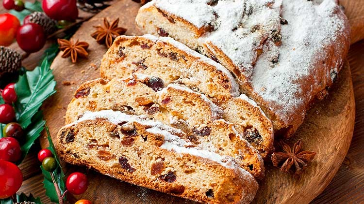 Angeschnittener, mit Puderzucker bestäubter Christstollen mit getrockneten Früchten auf einem Holzteller, daneben weihnachtliche Dekoration.