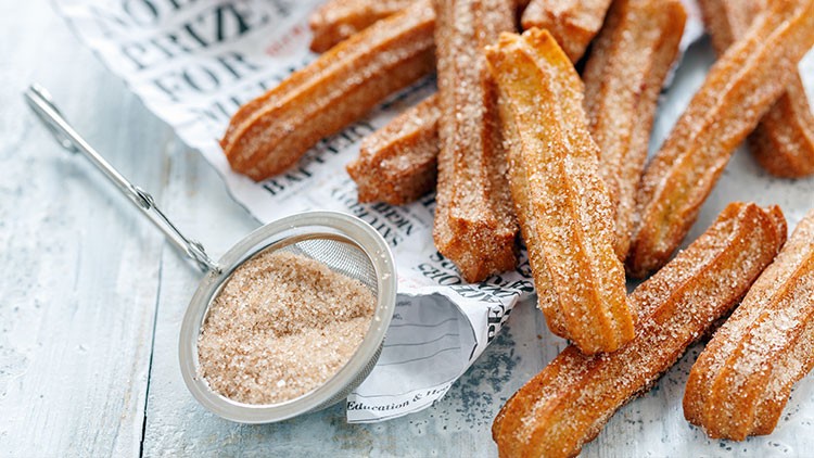 Mehrere mit Zucker bestreute Churros liegen auf Papier, daneben ein Sieb mit Zimt-Zucker-Mischung auf hellem Untergrund.