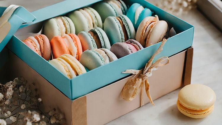 Eine türkisfarbene Schachtel mit bunten Macarons steht auf hellem Untergrund, daneben liegt ein einzelner gelber Macaron.
