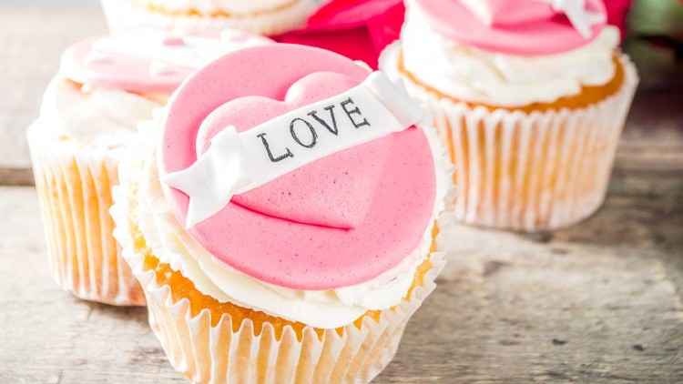 Vier Cupcakes mit weißem Frosting, rosa Herzdekor und dem Schriftzug LOVE auf einer Holzoberfläche, dahinter rote Rosen.