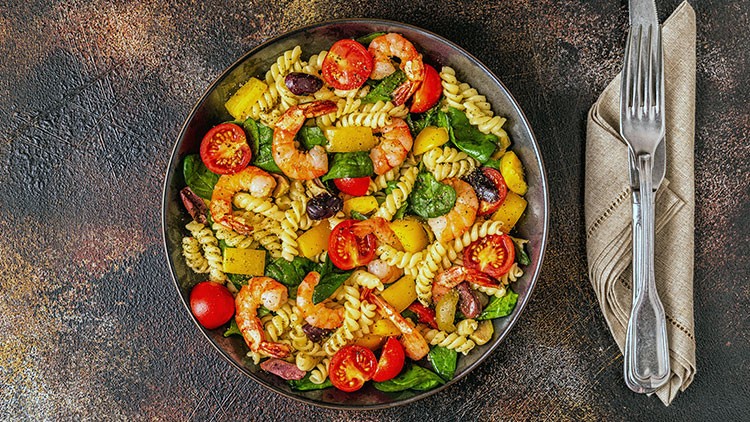 Ein Teller mit Nudelsalat aus Fusilli, Garnelen, Tomaten, Paprika, Oliven und Blattspinat, daneben Besteck auf einer Stoffserviette.
