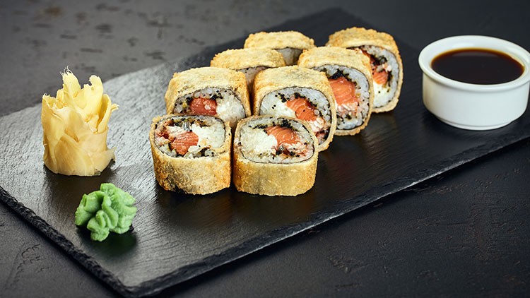 Acht frittierte Sushirollen mit Lachsfüllung auf schwarzer Platte, daneben Ingwer, Wasabi und eine weiße Schale mit Sojasoße.