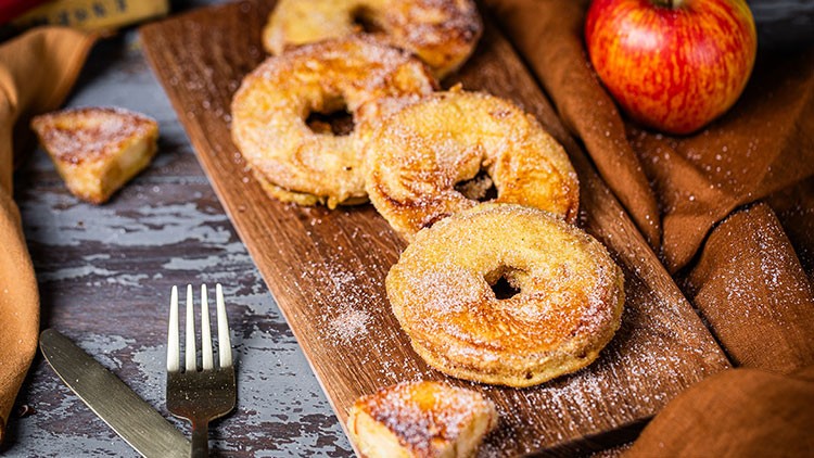 Mehrere gebackene Apfelringe mit Zucker auf einem Holzbrett, daneben ein roter Apfel, Besteck und ein braunes Tuch.