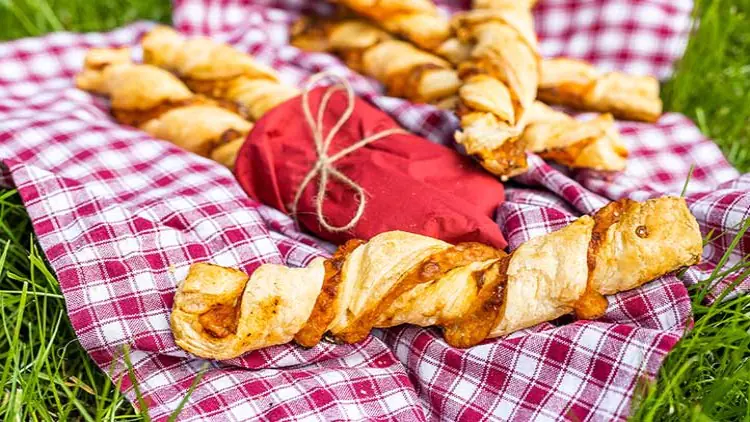 Blätterteig-Stangen mit Käse und Tomaten auf kariertem Tuch, daneben ein rotes Stoffbündel mit Kordel auf einer Wiese.