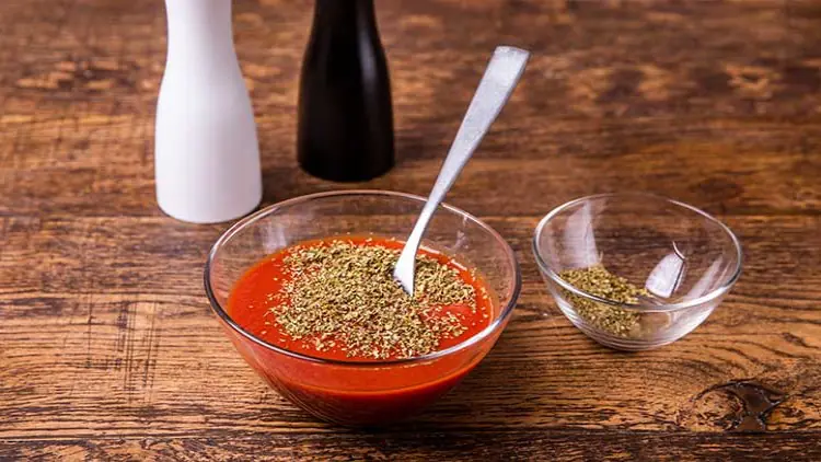 Eine Glasschüssel mit roter Tomatensoße, bestreut mit Kräutern, steht auf Holztisch, daneben Gewürzgläser und zwei Pfeffermühlen.