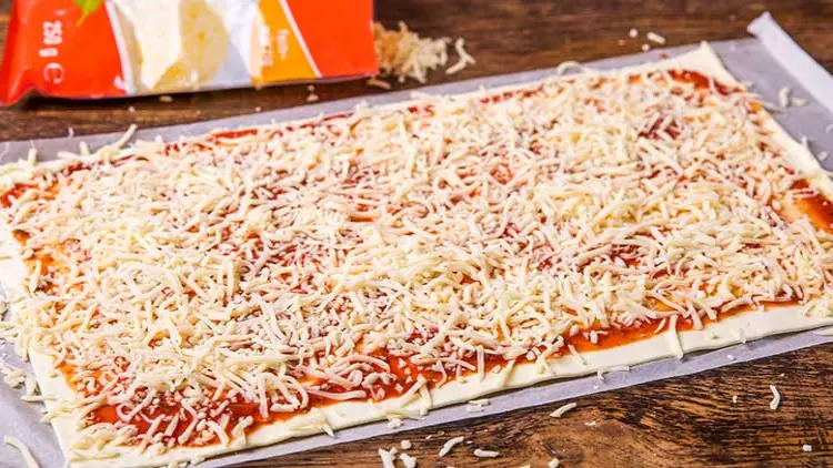 Ein rechteckiger ausgerollter Pizzateig mit Tomatensoße und geriebenem Käse, im Hintergrund ist eine angebrochene Käsepackung.