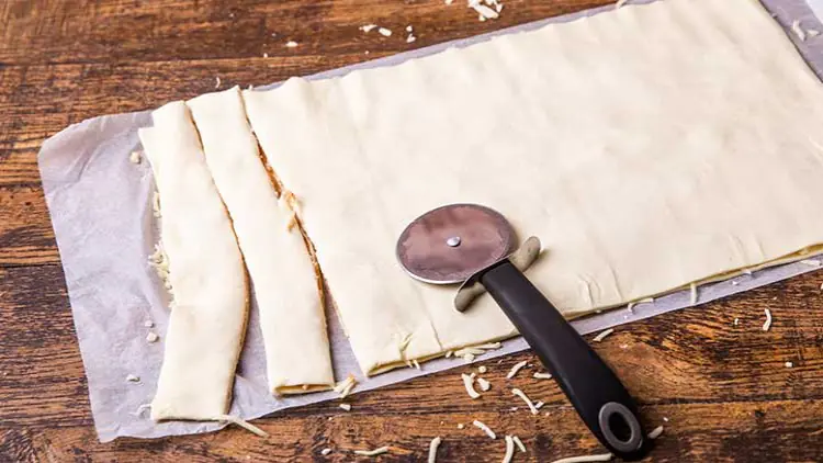 Ein rechteckiger ausgerollter Teig auf Backpapier mit abgetrennten Streifen und einem Pizzaschneider aus Metall und Kunststoff.
