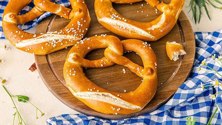 Drei goldbraune Brezeln mit Salz liegen auf einem runden Holzbrett, daneben ein blau-weiß kariertes Tuch und Wiesenblumen.