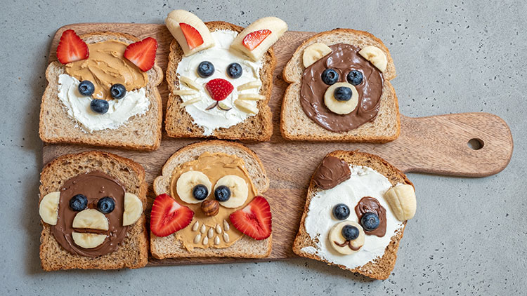Sechs Toastscheiben mit Aufstrichen und Früchten, darauf Tiergesichter aus Bananen, Erdbeeren, Blaubeeren und Nüssen.