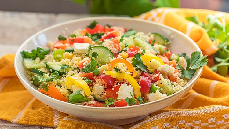 Ein Teller mit bunter Couscous-Salatmischung, Gurken, Paprika, Tomaten, Feta und frischer Petersilie auf einem gelben Tuch.