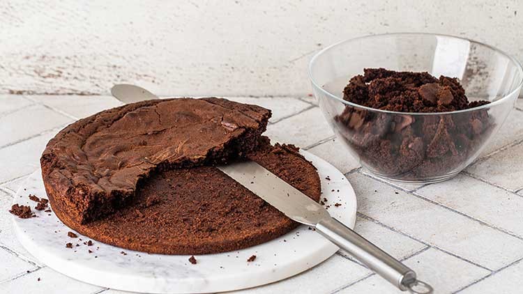 Ein angeschnittener Schokoladenkuchen liegt auf einem weißen Teller, daneben ein Messer und eine Glasschüssel mit Kuchenkrümeln.