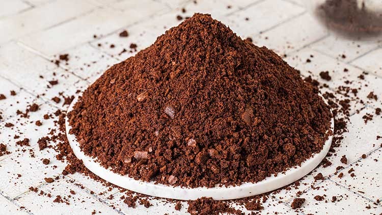 Ein Haufen braunes, gemahlenes Kaffeepulver liegt auf einem runden, weißen Untersetzer auf einem strukturierten Untergrund.