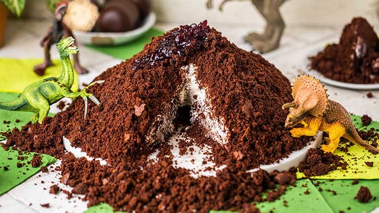 Ein aufgeschnittener Schokoladenkuchen in Form eines Hügels mit Cremefüllung und Deko-Dinosauriern auf grün-gelbem Untergrund.