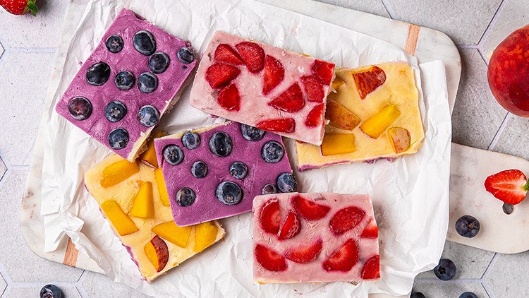 Sechs gefrorene Joghurtstücke mit Früchten wie Erdbeeren, Blaubeeren und Pfirsichen auf Backpapier und einem Holzbrett.