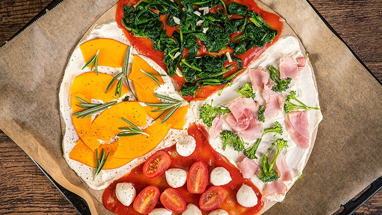 Runde Pizza auf Backpapier, belegt mit Spinat, Schinken, Hokkaidokürbis, Rosmarin, Tomaten, Mozzarella, Brokkoli und Tomatensauce.