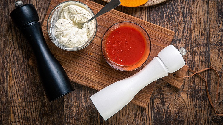 Schwarze und weiße Pfeffermühle mit Glasbehältern für Gewürzquark und rote Sauce auf einem Holzbrett, Holzuntergrund.