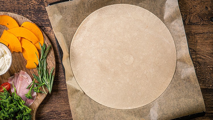 Ein runder Pizzateig liegt auf Backpapier, daneben ein Holzbrett mit Kürbis, Kräutern, Schinken, Tomaten und Frischkäse.
