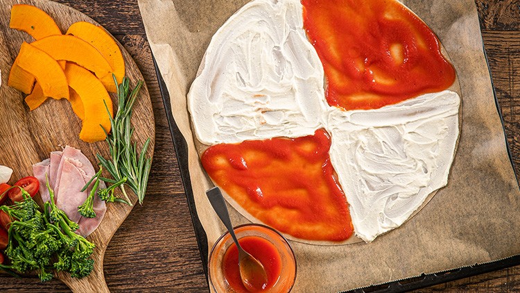 Flammkuchen mit weißem Belag und Tomatensoße wird vorbereitet, daneben liegen Gemüse, Kräuter, Schinken und Tomaten.