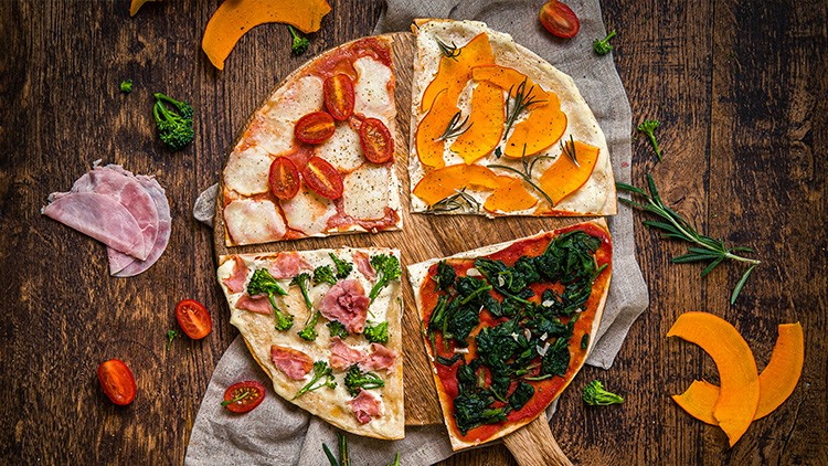 Vier Pizzastücke mit unterschiedlichen Belägen wie Tomate, Kürbis, Spinat und Schinken liegen auf einem Holztisch mit Deko.