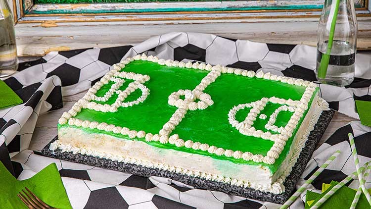 Rechteckige Torte mit grünem Belag und Sahneverzierungen in Form eines Fußballfeldes auf Tisch mit Fußballmuster-Dekoration.