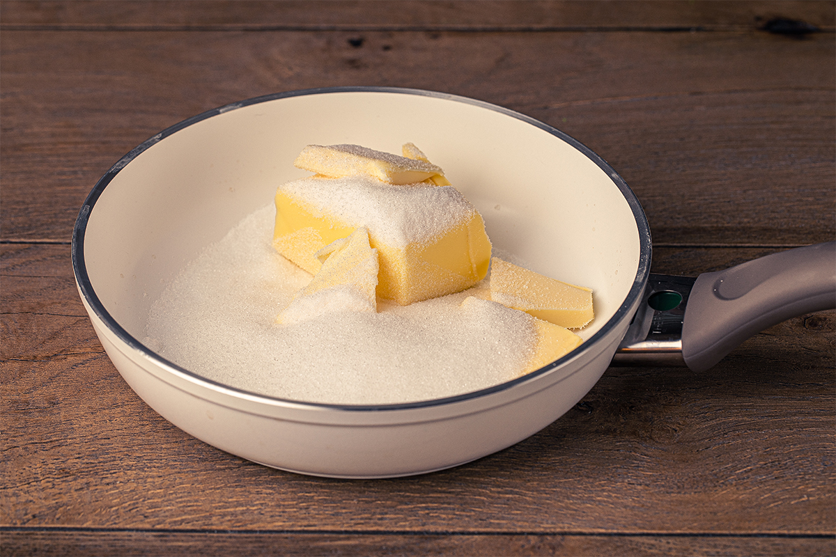Weiße Pfanne mit Butterstücken und weißem Zucker auf einer Holzoberfläche.