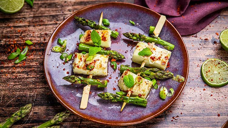 Gegrillte Halloumi-Spieße mit grünem Spargel, Minze, Frühlingszwiebeln und Chiliflocken auf einem violetten Teller.