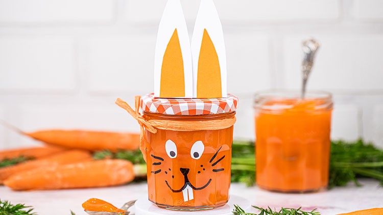 Ein Glas mit oranger Marmelade, verziert mit Hasengesicht und Ohren, steht vor frischen Karotten und einem weiteren Glas.