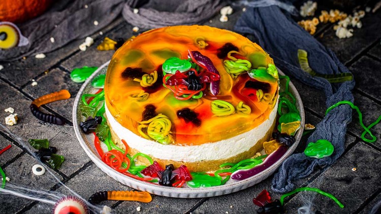 Runder Kuchen mit orangefarbener Geleeglasur, bunten Gummitieren dekoriert, auf einem grauen Teller, umgeben von Gummischlangen und Dekoration.
