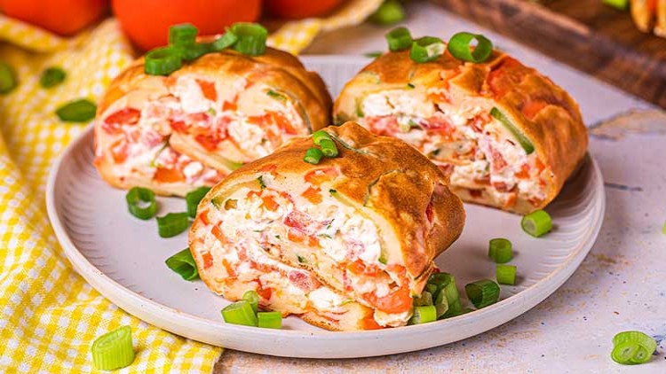 Drei goldbraune Pfannkuchenrollen mit Füllung aus Frischkäse, Tomaten und Kräutern auf einem Teller mit Frühlingszwiebeln.