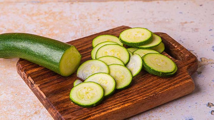 Eine frische Zucchini liegt neben mehreren gleichmäßig geschnittenen Zucchinischeiben auf einem hölzernen Schneidebrett.