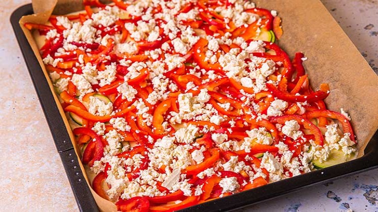 Ein mit Backpapier ausgelegtes Blech, belegt mit Zucchinischeiben, Paprikastreifen und zerbröseltem Feta.