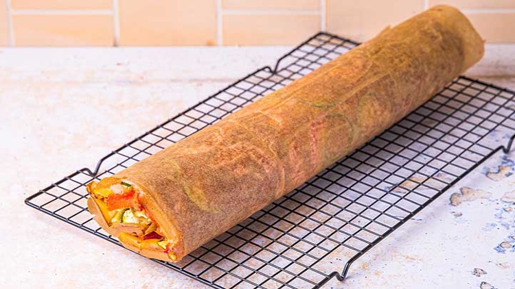Gerollter Wrap mit sichtbarer Füllung aus Gemüse und Käse liegt auf einem schwarzen Abkühlgitter vor einer beigen Kachelwand.