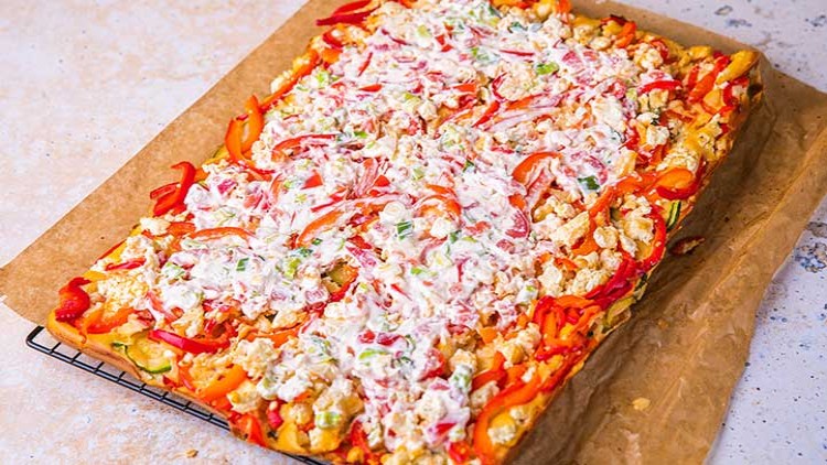 Rechteckiger Blechkuchen mit bunten Paprikastreifen, vegetarischen Zutaten und einer Creme auf Backpapier, auf einem Gitter liegend.