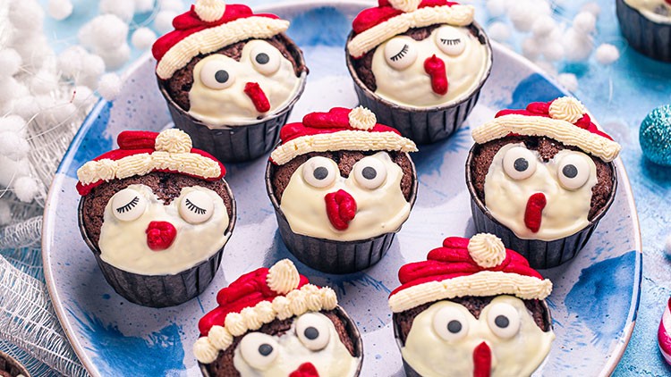 Sechs Schokomuffins mit Dekoration aus Creme und rotem Zuckerguss, gestaltet als Weihnachtsgesichter mit Mützen.
