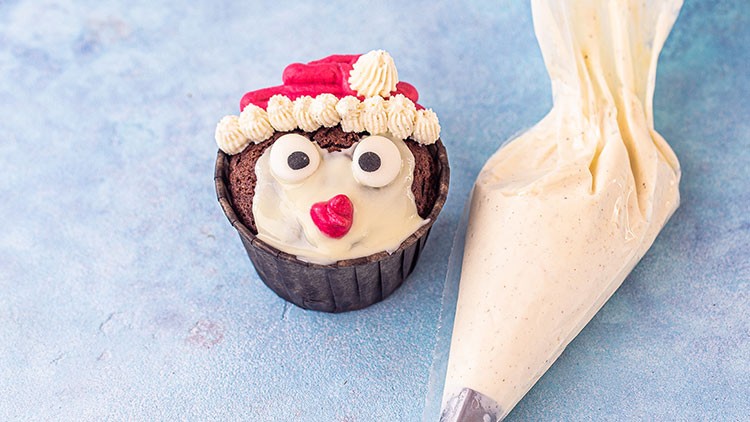 Ein Schokoladen-Muffin mit Zuckerdekoration als Gesicht und Zipfelmütze neben einem Spritzbeutel mit heller Creme.