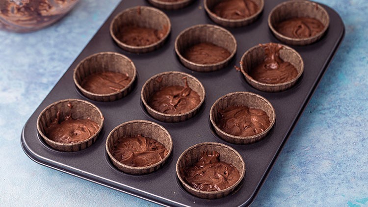 Eine Muffinform mit zwölf Mulden, gefüllt mit braunem Teig in papierartigen Förmchen, steht auf einer hellblauen Fläche.