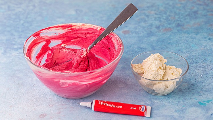 Eine Glasschüssel mit rosa Creme, daneben eine kleine Glasschüssel mit weißer Creme und eine Tube Speisefarbe Rot.