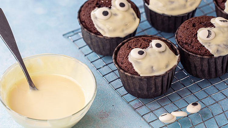 Schokoladenmuffins mit weißem Zuckerguss und Zuckeraugen stehen auf einem Gitterrost, daneben eine Schüssel mit Zuckerguss.