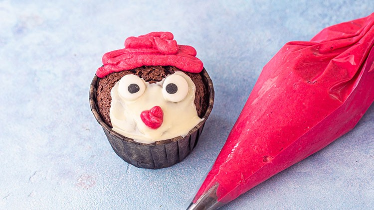 Schokoladenmuffin mit Augen aus Zuckerguss und rotem Zuckerdekor neben einer Spritztüte mit roter Creme auf blauem Untergrund.