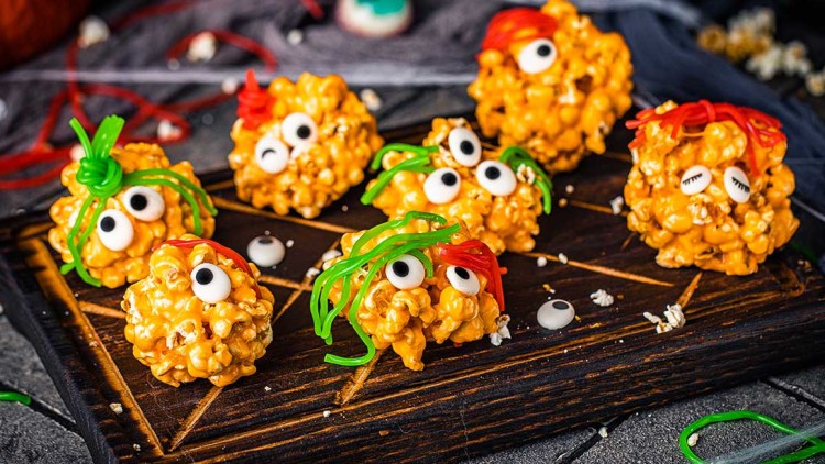 Orangene Popcorn-Bälle mit Zuckerschrift, Zuckeraugen und grünen oder roten Süßigkeitenfäden auf einem Holzbrett.