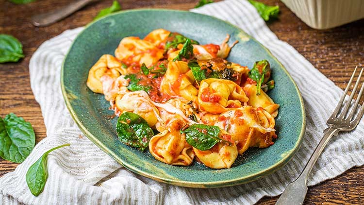 Gefüllte Tortellini in Tomatensauce mit frischem Babyspinat auf grünem Teller, daneben eine Gabel und ein Leinentuch.